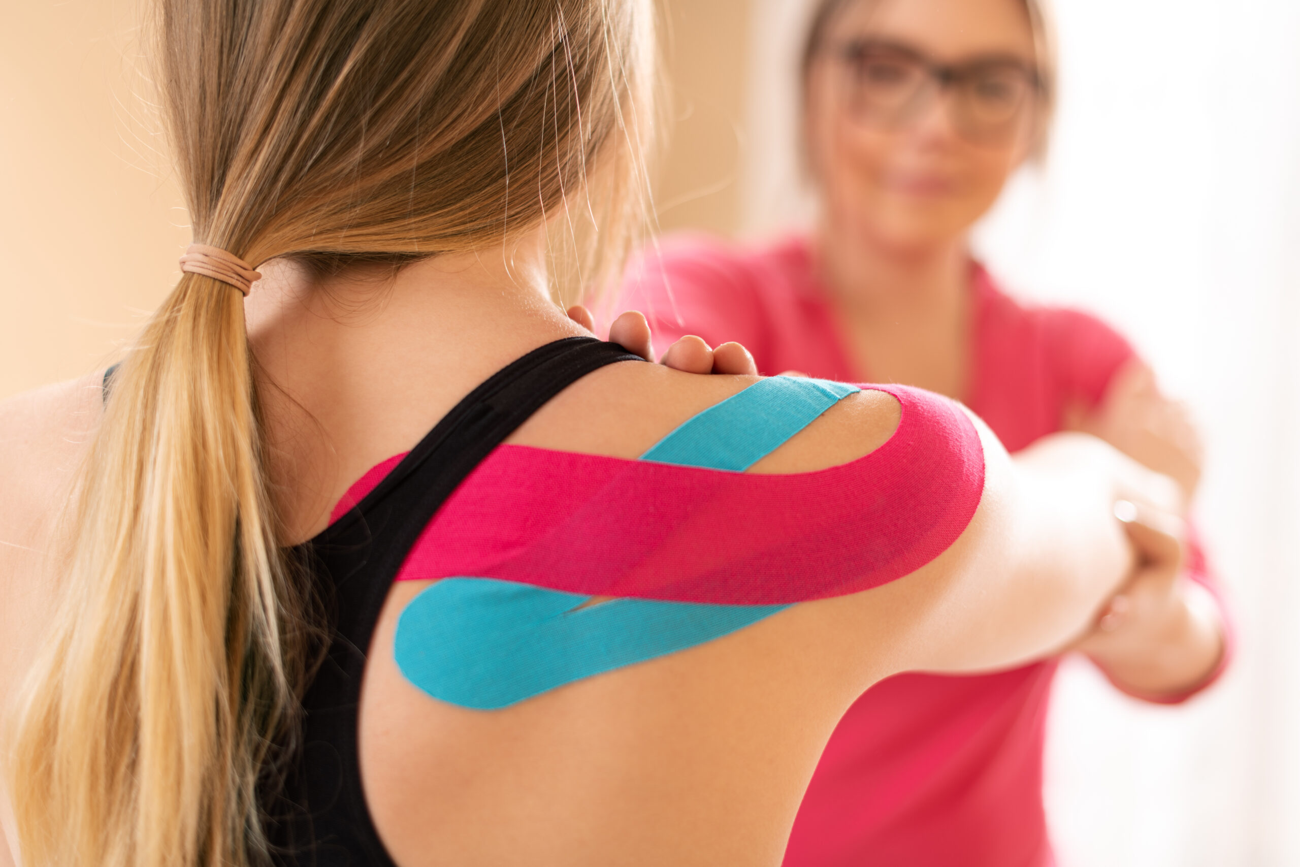 Young female patient wearing kinesio tape on her shoulder exercising with a professional physical therapist. Kinesiology, physical therapy, rehabilitation concept. Stretching Klaff Sports Physical Therapy